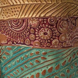 May include: Close-up of a ceramic vase with a multi-colored, textured design. The vase features a brown top with a geometric pattern, a burgundy middle section with a swirling pattern, and a teal bottom with a wavy pattern.