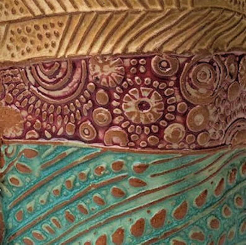 May include: Close-up of a ceramic vase with a multi-colored, textured design. The vase features a brown top with a geometric pattern, a burgundy middle section with a swirling pattern, and a teal bottom with a wavy pattern.