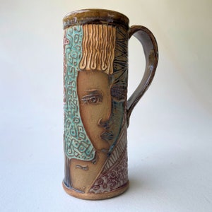 May include: A ceramic mug with a brown, blue, and white design featuring a stylized face. The mug has a handle and is made in a rustic style.