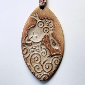 Pode incluir: Pingente oval de cerâmica com um desenho de ovelha. O pingente é bege com uma imagem de ovelha mais clara e detalhes em marrom. A ovelha tem olhos grandes e lã encaracolada. Uma fita marrom está presa para pendurar.