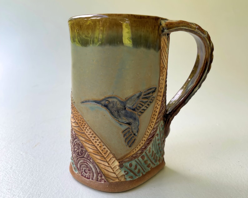 May include: A ceramic mug with a green, brown, and blue glaze. The mug has a handle and a hummingbird design on the front.