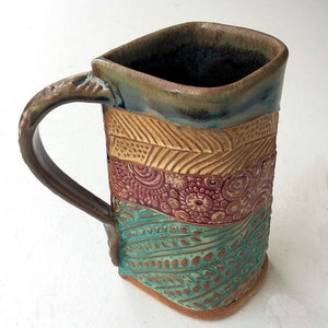 May include: A handmade ceramic mug with a square body and a handle. The mug is decorated with a multi-colored pattern of brown, yellow, red, and blue.