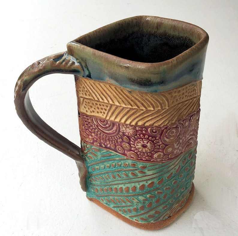 May include: A handmade ceramic mug with a square body and a handle. The mug is decorated with a multi-colored pattern of brown, yellow, red, and blue.