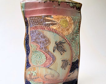 May include: A ceramic vase with a textured surface and a colourful design featuring a sun, palm trees, and sea turtles. The vase is a light brown colour with shades of blue, green, purple, and red.