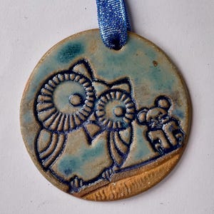 May include: A round ceramic ornament with a blue ribbon. The ornament features a relief design of two owls and a small animal, in shades of blue and brown. The ornament is approximately 3 inches in diameter.