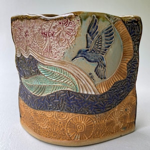 Puede incluir: Un recipiente de cerámica cilíndrico que presenta un colibrí tallado en relieve en pleno vuelo sobre un fondo circular. La superficie muestra texturas intrincadas, incluyendo patrones florales en marrón rojizo, hojas verdes y una banda azul oscuro.