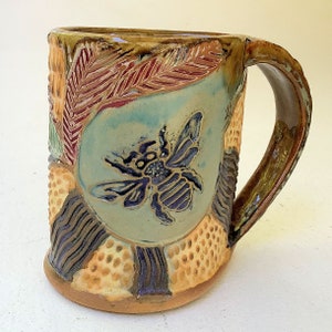 Könnte beinhalten: Eine Keramiktasse mit einem Bienendesign. Die Tasse ist mit verschiedenen Farben und Texturen verziert, darunter Braun, Gelb, Grün, Blau und Lila. Die Biene befindet sich in der Mitte der Tasse und ist von einem Wabenmuster umgeben.