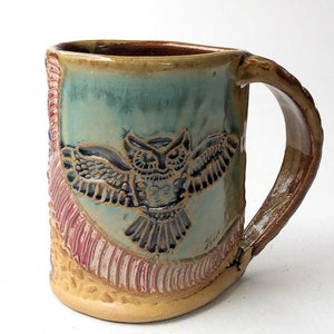 May include: A ceramic mug with a light blue glaze and a brown handle. The mug features a raised design of an owl with its wings spread. The owl is in shades of brown, black, and blue. The mug also has a decorative band of pink and red stripes around the bottom.