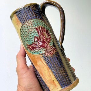 May include: A tall, handcrafted ceramic mug with a large handle. The mug features a detailed relief of a bird in flight, set against a textured green background. The mug's exterior is adorned with vertical and geometric patterns in shades of blue, orange, and brown.