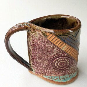 May include: A handmade ceramic mug with a brown, orange, and purple geometric design. The mug has a unique, textured surface and a large handle.