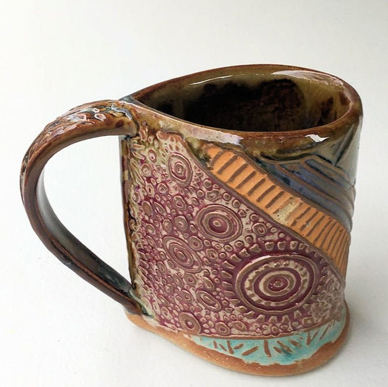 May include: A handmade ceramic mug with a brown, orange, and purple geometric design. The mug has a unique, textured surface and a large handle.