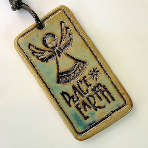 May include: A ceramic ornament with a blue-green glaze features a relief image of an angel with the words "Peace on Earth" in black lettering.