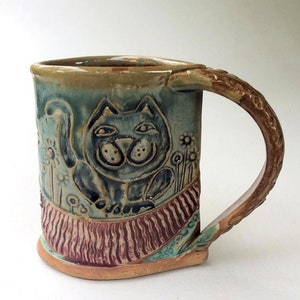May include: A handmade ceramic mug with a whimsical design featuring a cat with a striped tail and flowers. The mug has a brown handle and a green, brown, and red glaze.