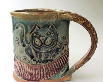 Może przedstawiać: Ręcznie robiony kubek ceramiczny z fantazyjnym wzorem przedstawiającym kota z paskowanym ogonem i kwiatami. Kubek ma brązowy uchwyt i zieloną, brązową i czerwoną glazurę.