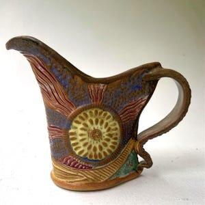 May include: A ceramic pitcher featuring a prominent central yellow circular motif with radiating lines. The surface is decorated with textured blue, red, and tan patterns, including a curved handle and a wide pouring spout with a brown clay base.
