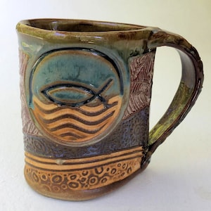 May include: A handmade ceramic mug with a brown, green, and blue glaze. The mug features a raised design of a fish and waves in a circle. The mug has a textured handle and a decorative base.