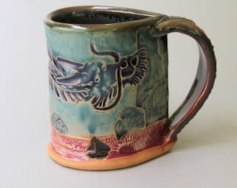 May include: A ceramic mug with a blue and green glaze. The mug has a handle and a dragon design on the front. The mug is sitting on a white surface.