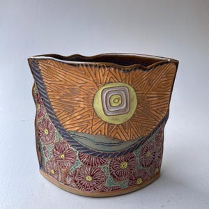 May include: A hand-built ceramic vessel with an irregular, wavy rim. The upper section features a carved sun motif with radiating lines in shades of orange and yellow. The lower portion displays a repeating pattern of textured, dark pink flowers.