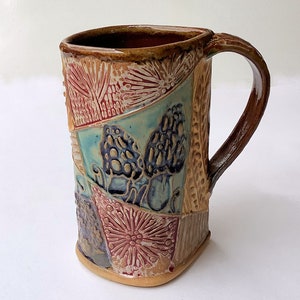 Può includere: Una tazza in ceramica fatta a mano con un design a patchwork che presenta funghi, fiori e motivi geometrici in tonalità di marrone, blu, verde e rosso.