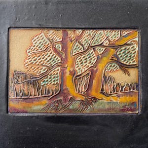 May include: A framed ceramic tile featuring a detailed relief of a tree with a yellow trunk and branches, set against a tan background. The artwork includes a depiction of a forest and is encased in a black frame.