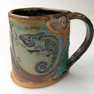 May include: A ceramic mug with a brown, green, and blue glaze. The mug features a raised design of a chameleon with a spiral shell. The mug has a handle and is signed "3E..." on the side.