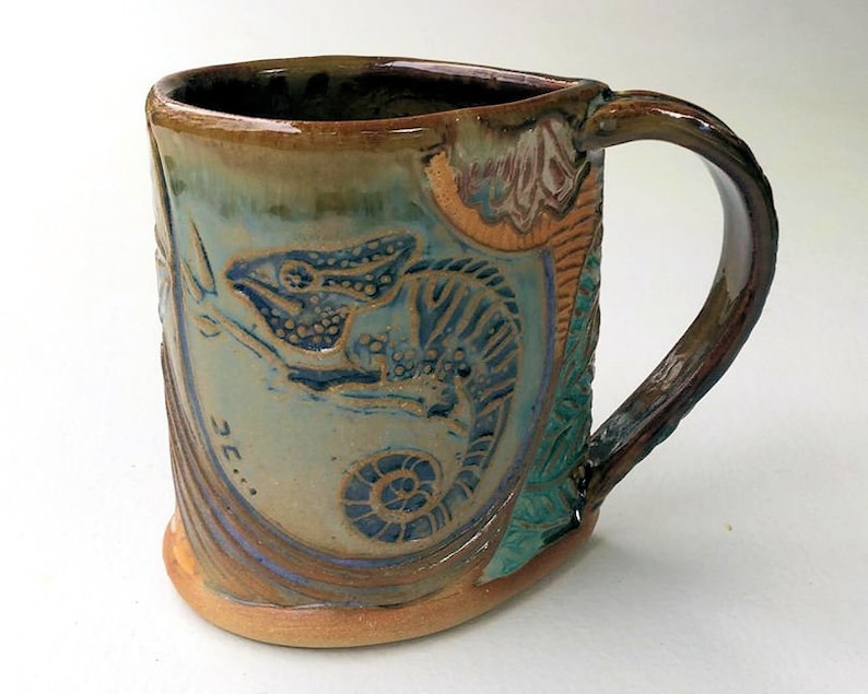 May include: A ceramic mug with a brown, green, and blue glaze. The mug features a raised design of a chameleon with a spiral shell. The mug has a handle and is signed "3E..." on the side.