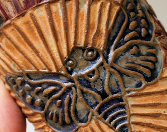 May include: A close-up of a ceramic mug featuring an intricate, embossed design of a moth with brown, blue and white accents. The moth is positioned centrally against a backdrop of radiating lines in a warm brown colour.