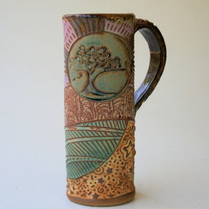 May include: A handmade ceramic mug featuring a tree design within a circle, surrounded by a patterned design in shades of brown, pink, and green. The mug has a handle and is crafted in a rustic style.