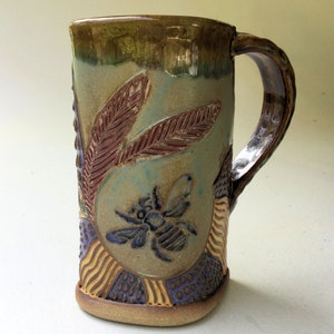 May include: A handmade ceramic mug with a green, brown, and purple glaze. The mug features a bee and leaf design.