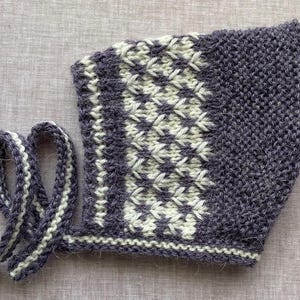May include: A knitted bonnet featuring a dark purple and cream color scheme. The main body displays a textured garter stitch pattern, while a central band showcases a detailed star-like geometric motif. Two long knitted ties extend from the base.