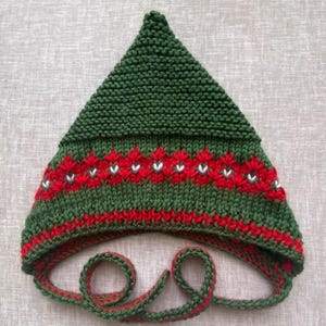 May include: A pointed green knit hat featuring a horizontal band of red and white floral-inspired patterns. The hat is constructed with a garter stitch texture and includes two long, integrated green and red braided ties at the base.