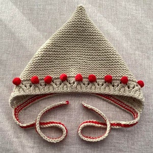 May include: A beige knitted baby bonnet with red pom-poms around the brim and red and white striped ties.