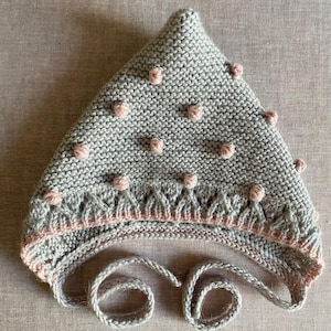 May include: A gray knitted hat with pink accents and pom-poms. The hat features a decorative pattern along the bottom edge and ties with two long, knitted strings. The hat is designed for warmth and style.