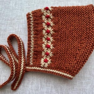 May include: A hand-knitted bonnet in a rich rust-orange color featuring a garter stitch texture. The design includes a vertical band of cream-colored embroidery with small, bright red pom-poms and matching cream and rust-orange braided ties.
