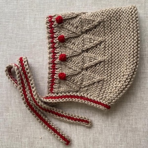 May include: A knitted bonnet in a light tan color with red accents. The bonnet features a textured pattern on the crown and is trimmed with red yarn. Three small red pom-poms are sewn on the side. Red ties are attached to the bonnet.
