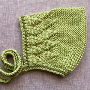 May include: A green knitted baby bonnet with a cable knit pattern and a long tie.