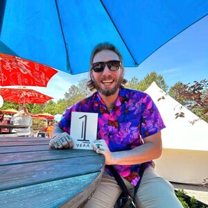 May include: A man wearing sunglasses and a purple and pink floral shirt is sitting at a wooden table under a blue umbrella. He is holding a white card with the number "1" and the word "year" written on it.
