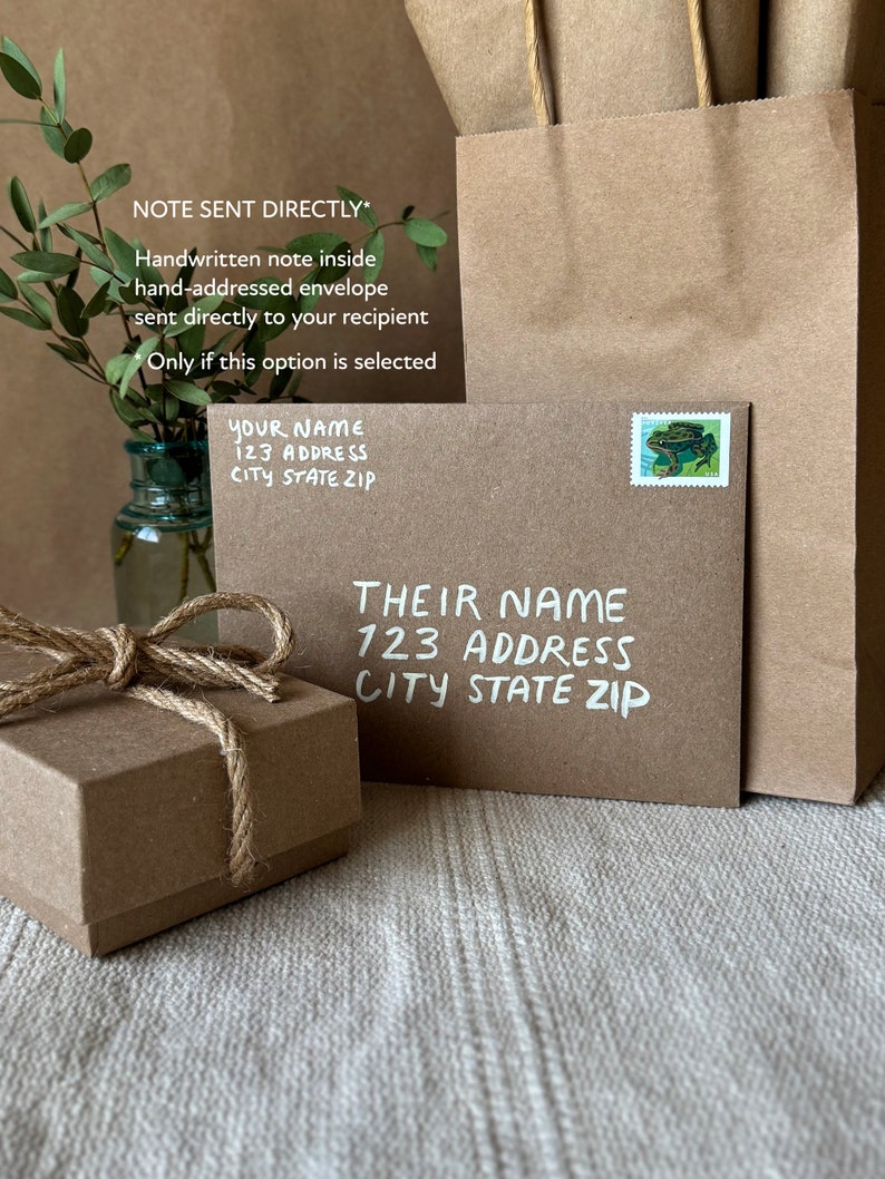 May include: A brown paper envelope with a handwritten note inside. The envelope is addressed to "YOUR NAME 123 ADDRESS CITY STATE ZIP" and has a stamp with a green frog on it. The envelope is sitting on a white surface next to a small brown box with a twine bow.