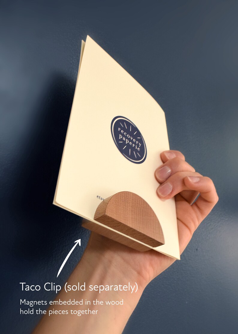 May include: A wooden taco clip with magnets embedded in the wood to hold the pieces together. The clip is holding a card with the text "recovery paperie" on it.