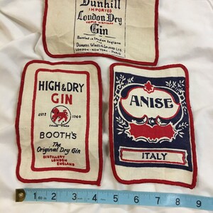 May include: Three vintage fabric patches with red stitching. The top patch is white with black text that reads "Dunhill Imported London Dry Gin Triple Distilled Bottled in London England Dunhill Wines & Spirits Ltd. London - New York - Paris". The middle patch is white with red and blue text that reads "High & Dry Gin 2572 Trade Mark 1740 Booth's The Original Dry Gin Distillery London England". The bottom patch is white with red and blue text that reads "Anise Italy".