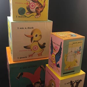May include: A stack of colorful wooden blocks with animal illustrations and text. The top block features a cat with the text "I am a cat, I meow". The second block features a duck with the text "I am a duck, I quack". The third block features a bird with the text "I am a bird, I sing". The fourth block features a monkey with the text "I do tricks". The bottom block features a dog with the text "I am a dog, I bark".