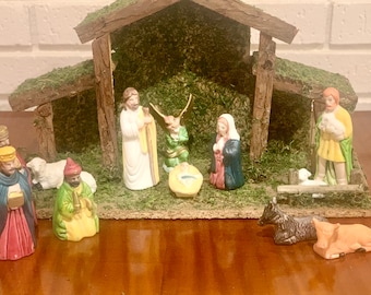 Pode incluir: Um presépio tradicional com um estábulo de madeira, musgo e figuras de Maria, José, o menino Jesus, um pastor, um anjo e três reis magos. As figuras são de cerâmica e pintadas em cores vibrantes.