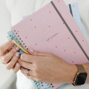 A model is holding two ivf journey planners and one fet journey planner, the pink spot cover design is the most visible with the mint pineapple one behind it and the blue star fet planner at the bottom.