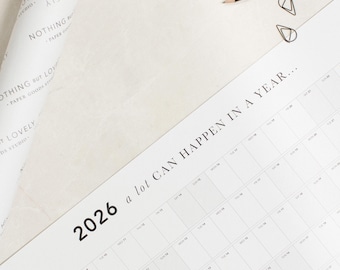 May include: A white calendar with the text "2026 a lot can happen in a year..." printed on it. The calendar is on a white surface with a roll of wrapping paper and two paper clips.