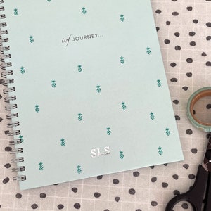 May include: A light blue spiral notebook with a pineapple pattern and the text "iuf JOURNEY..." and "SLS" on the cover.