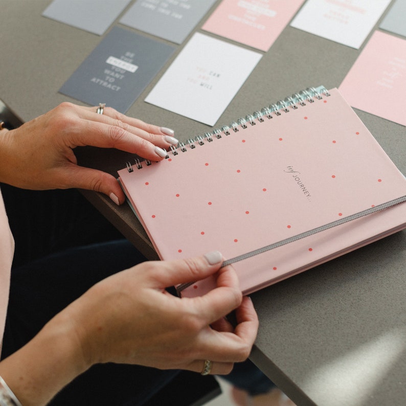 A model is holding the pink spot ivf planner design, she is about to open it removing the grey elastic closure. There are two week wait support cards on the table underneath the planner.