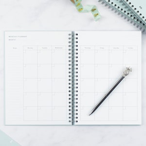 The ivf journey planner open on a table showing a double page spread of a monthly calendar page. A pineapple shaped biro lays on top of one of the pages.