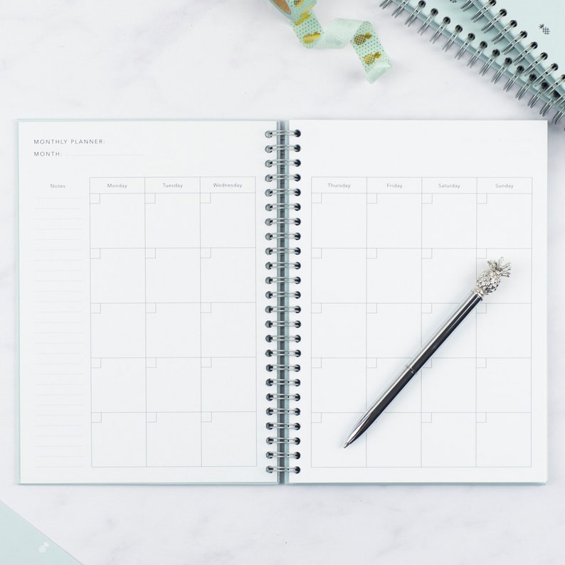 The ivf journey planner open on a table showing a double page spread of a monthly calendar page. A pineapple shaped biro lays on top of one of the pages.