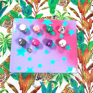 Peut inclure: Un plateau rectangulaire violet avec un dégradé rose et des étoiles turquoise. Le plateau contient huit boutons colorés de différentes couleurs, dont blanc, rose, vert et violet. Un petit objet argenté est également présent. Le fond présente un imprimé tropical.