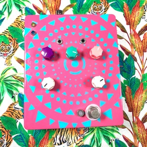 May include: A pink effects pedal with a geometric design in turquoise. It features six colored knobs in purple, green, and pink, and two white knobs. The pedal has a silver footswitch and is set against a tropical print background.